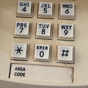 May include: Close-up of a cream-colored telephone keypad. The keypad features white square buttons with black numbers and letters. The buttons include numbers 0-9, the asterisk, and the pound sign. The text "AREA CODE" is also visible.