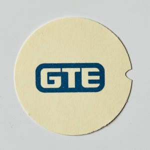 May include: A round, off-white paper coaster with a dark blue rectangular logo that reads "GTE". The coaster has a small notch cut out of the edge.
