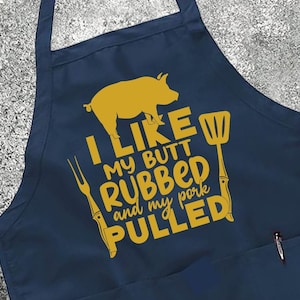 May include: Navy blue apron with a gold pig graphic and the text "I LIKE MY BUTT RUBBED and my PORK PULLED." The apron has a pocket and adjustable neck strap. The apron is surrounded by food items like tomatoes, pasta, and olives.