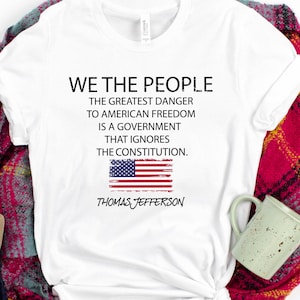 May include: White t-shirt with the text "WE THE PEOPLE" and a quote by Thomas Jefferson. The shirt is paired with blue jeans, tan sandals, and a red and black plaid scarf. A decorative acorn and a green mug are also present.