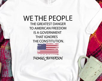 以下が含まれることがあります： 「WE THE PEOPLE」の文字とトーマス・ジェファーソンの引用句が書かれた白いTシャツ。青いジーンズ、タン色のサンダル、赤と黒のチェック柄のスカーフと組み合わせています。装飾的なドングリと緑色のマグカップも含まれています。