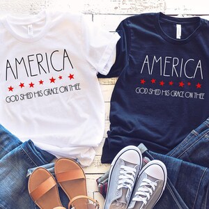May include: Two t-shirts, one white and one navy blue, with the word "AMERICA" and the phrase "GOD SHED HIS GRACE ON THEE" printed on them. The shirts are paired with blue jeans, sandals, and sneakers, creating a patriotic theme.