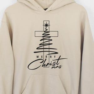 May include: A beige hoodie with a front pocket and a drawstring hood. The front features a black graphic of a cross and Christmas tree design with the words "Merry Christ mas" in a script font.