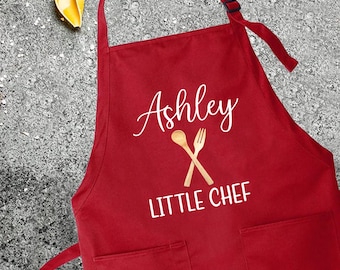 May include: Red apron with the name "Ashley" and the text "Little Chef" printed in white. The apron has two pockets and a white graphic of a wooden spoon and fork.