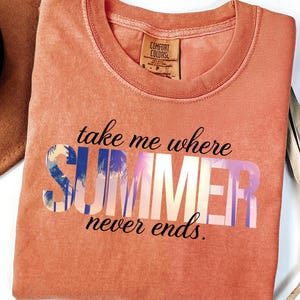 May include: Coral-colored sweatshirt with the text "take me where SUMMER never ends" in a gradient font. A brown hat with a black band is in the upper left corner. A dried palm leaf is on the right.