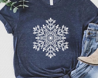 May include: A navy blue t-shirt featuring a large white snowflake design. The shirt is paired with blue jeans and tan ankle boots. The image suggests a winter or holiday theme.