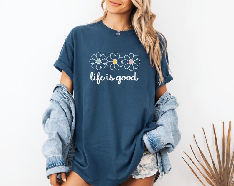 May include: A navy blue t-shirt with three daisy flower graphics in white, yellow, and pink. The words "life is good" are written in white script below the flowers. The shirt is worn with a light blue denim jacket.