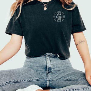 May include: A black t-shirt with a white circle on the front that says "Your Logo Here". The shirt is being worn by a person with long blonde hair and blue jeans.