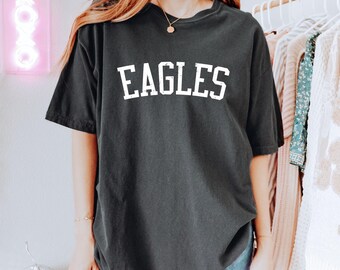 May include: Dark gray t-shirt with the word "EAGLES" printed in white block letters. The shirt is a short-sleeved, crew neck style. The model is wearing blue jeans. A neon sign that says "XOXO" is in the background.
