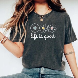 May include: Dark gray t-shirt with the phrase "life is good" in white script. Above the text are three daisy flowers with pastel blue, yellow, and pink centers. The shirt is a casual, comfortable style.