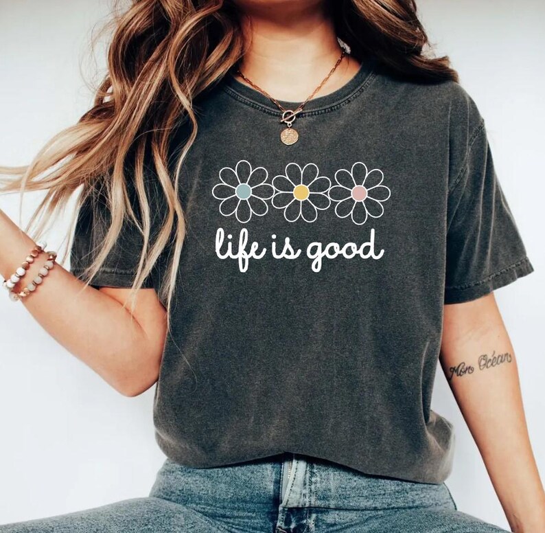 May include: Dark gray t-shirt with the phrase "life is good" in white script. Above the text are three daisy flowers with pastel blue, yellow, and pink centers. The shirt is a casual, comfortable style.