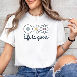 May include: White t-shirt with a daisy graphic and the phrase "life is good" in a handwritten font. The daisy graphic features three flowers with pastel blue, yellow, and pink centers. The shirt is paired with blue denim jeans.