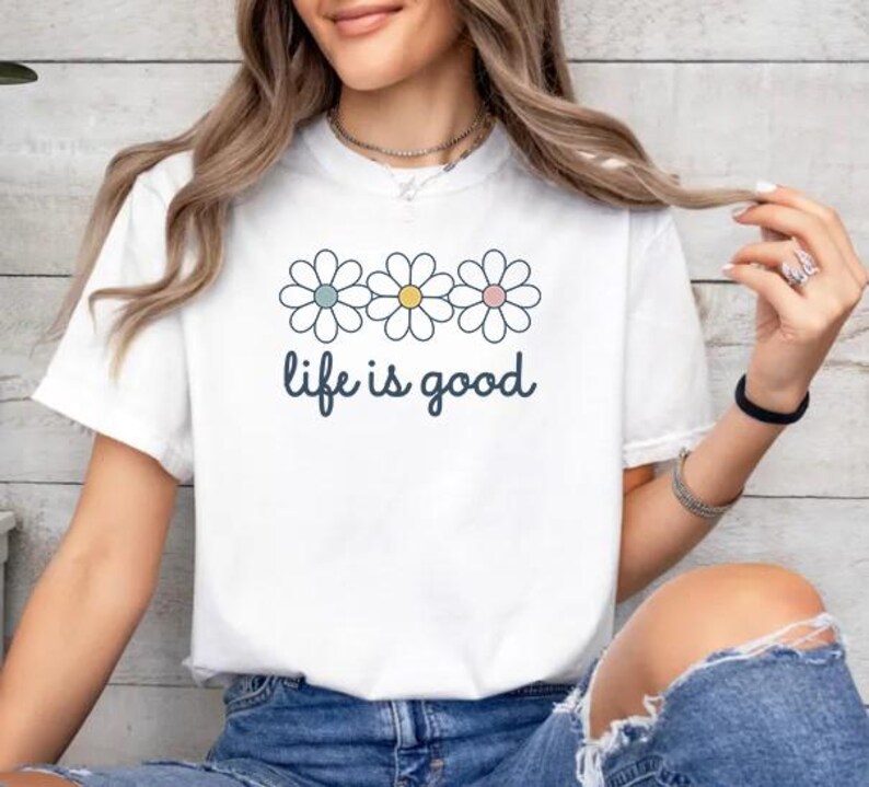 May include: White t-shirt with a daisy graphic and the phrase "life is good" in a handwritten font. The daisy graphic features three flowers with pastel blue, yellow, and pink centers. The shirt is paired with blue denim jeans.