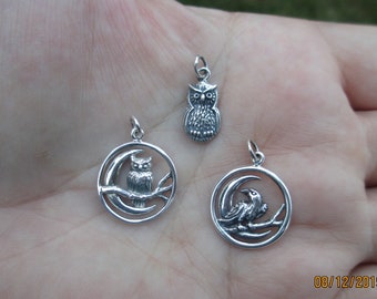 May include: Three silver charms featuring owls and a raven. The charms are in the shape of a crescent moon with an owl or raven perched on a branch. The charms are on a hand.
