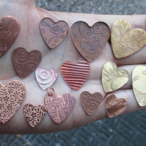 May include: A collection of 14 heart-shaped metal pendants with various designs and finishes, including copper, gold, and silver. Some pendants feature intricate patterns, while others have a more simple design. The pendants are arranged on a hand, showcasing their size and detail.