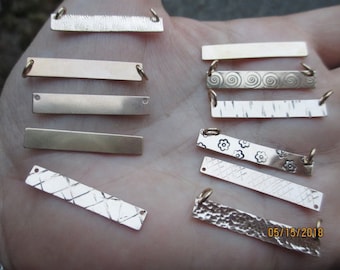 May include: A collection of ten gold-toned metal bar pendants with various textures and designs. Some pendants are plain, while others feature swirls, flowers, and a criss-cross pattern. The pendants are arranged on a hand.
