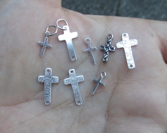 May include: A collection of seven silver cross charms with different designs. Some of the crosses have a swirl pattern, while others are plain. The charms are all about 1 inch in size.