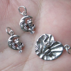 May include: Three silver charm pendants featuring frogs. One charm shows a frog sitting on a crescent moon, another shows a frog sitting on a lily pad, and the third charm shows a frog sitting on a crescent moon.