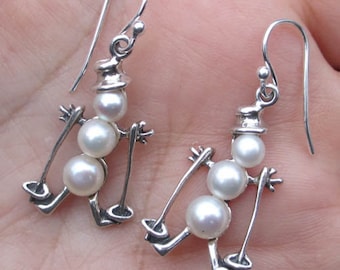 May include: Silver earrings featuring a snowman design with three white pearls. The snowman is holding ski poles.