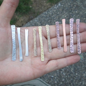 Puede incluir: Surtido de diez colgantes de barra de metal en varios acabados, incluyendo martillados, texturizados y estampados. Las barras son todas aproximadamente del mismo tamaño y forma, y están dispuestas en una fila en una mano.