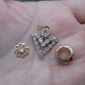 May include: Three gold-toned jewelry findings, including a flower-shaped piece with a clear rhinestone in the center, a round piece with a textured surface, and a pendant with a clear rhinestone cluster in a triangular shape.