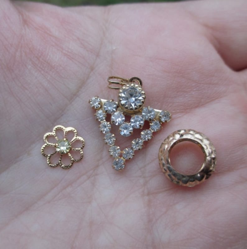 May include: Three gold-toned jewelry findings, including a flower-shaped piece with a clear rhinestone in the center, a round piece with a textured surface, and a pendant with a clear rhinestone cluster in a triangular shape.