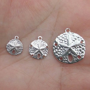 Op de afbeelding: Drie zilverkleurige zanddollarbedels van verschillende formaten. Elke bedel heeft een zeesterontwerp in het midden en kleine gaatjes aan de randen. De bedels hebben een lus aan de bovenkant om aan sieraden te bevestigen.