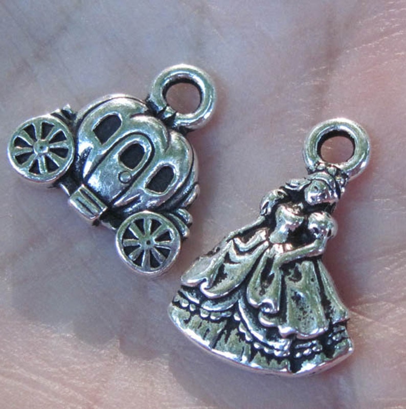 May include: Two silver charm pendants, one depicting a princess in a gown and the other a carriage. Both charms have a loop for attaching to a necklace or bracelet.
