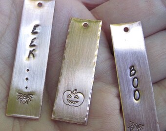 May include: Three copper-colored metal tags with different Halloween-themed designs. The first tag has the word "EEK" and a spider. The second tag has a pumpkin face. The third tag has the word "BOO" and a spider.