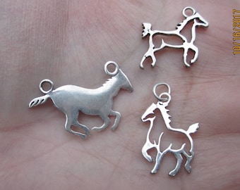 Peut inclure: Trois breloques de cheval en argent, chacune avec un design différent. Les breloques sont sur une main.