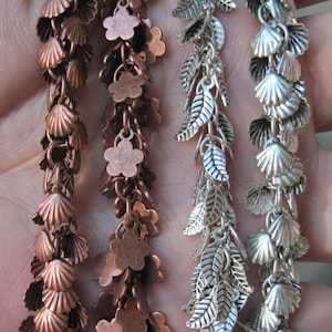 May include: Close-up of four strands of decorative chain. Two are copper-toned, featuring seashell and flower charms. The other two are silver-toned, with leaf and seashell charms. The chains are linked together, creating a cascading effect.