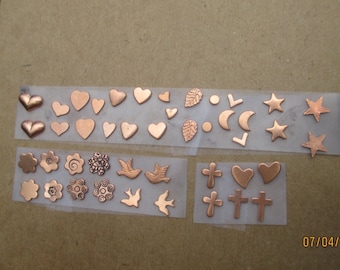May include: A sheet of copper charms with various designs, including hearts, leaves, stars, crosses, and birds. The charms are arranged in rows on a white background.