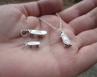 May include: Two silver skateboard charms, one on a chain necklace and one on a ring. The charms are detailed with wheels and a textured board.