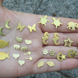 May include: A collection of gold-colored metal charms in various shapes, including stars, fish, butterflies, hearts, leaves, and animals. The charms are arranged on a hand.