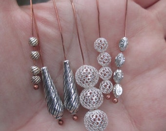 May include: Five silver and copper wire earrings with various silver beads and charms. The earrings feature a variety of shapes, including round, teardrop, and oval. The beads are all silver and have a textured finish. The charms are also silver and have a variety of designs.