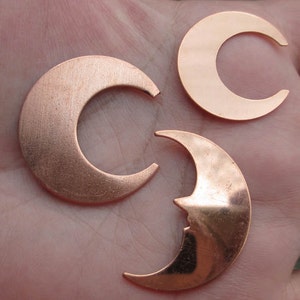 Puede incluir: Tres formas de media luna de cobre, cada una con un diseño diferente. Las lunas están sobre una superficie marrón claro.