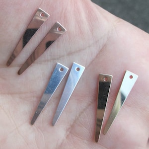 Puede incluir: Seis hallazgos de pendientes de metal en dos acabados diferentes. Dos son de color dorado y cuatro son de color plateado. Todos son de forma triangular con un pequeño agujero en la parte superior.