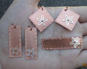 May include: Five copper-colored metal tags with a floral design. Each tag has a white flower with a small copper-colored center. The tags are arranged on a hand.