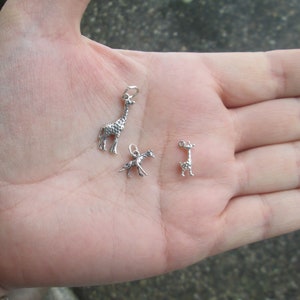 May include: Three silver giraffe charms, two are standing and one is laying down. The charms are on a person's hand.