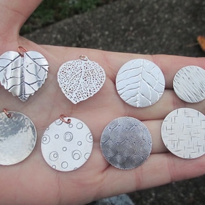 May include: Seven silver pendants with different designs, including leaves, circles, and geometric patterns. The pendants are displayed on a hand.