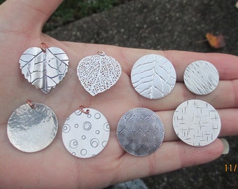 May include: Seven silver pendants with different designs, including leaves, circles, and geometric patterns. The pendants are displayed on a hand.