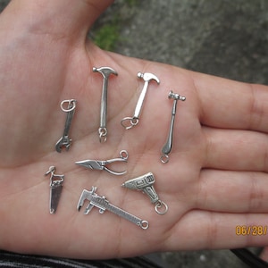 May include: A collection of silver charm pendants in the shape of tools, including a hammer, saw, pliers, wrench, and a drill. The charms are displayed on a hand.