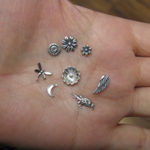 Puede incluir: Una colección de dijes de plata con un diseño de libélula, una luna creciente, un pájaro, un ala y tres diseños de flores diferentes.