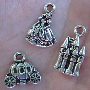 May include: Three silver charm pendants featuring a princess, a castle, and a pumpkin carriage. The princess charm is wearing a gown with a ruffled skirt. The castle charm has a pointed roof and multiple windows. The pumpkin carriage charm has a rounded shape with wheels.