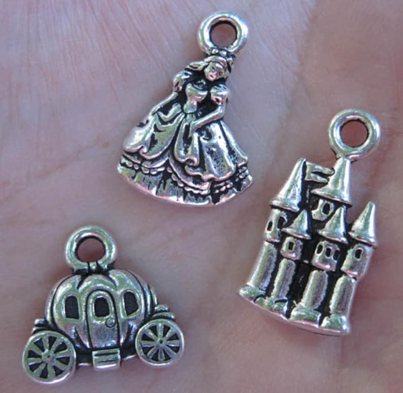 May include: Three silver charm pendants featuring a princess, a castle, and a pumpkin carriage. The princess charm is wearing a gown with a ruffled skirt. The castle charm has a pointed roof and multiple windows. The pumpkin carriage charm has a rounded shape with wheels.