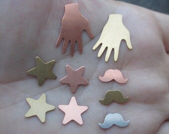May include: Assortment of metal charms in various shapes and colors, including stars, hands, and mustaches. The charms are made of copper, brass, and silver.