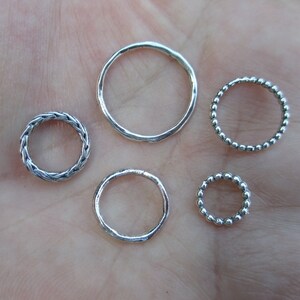 May include: Five silver rings of different styles and sizes. The rings are on a light brown surface.