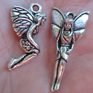 May include: Two silver charm pendants in the shape of fairies. Both fairies have wings and are standing with their arms at their sides. The fairy on the left is in a crouching position with her wings spread out behind her. The fairy on the right is standing straight up with her wings spread out behind her.