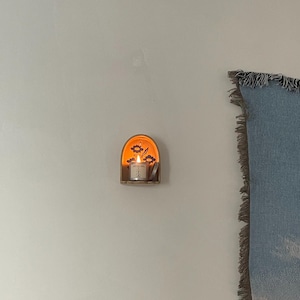 Pu&ograve; includere: Un piccolo portacandele da parete in legno con una candela accesa. Il portacandele presenta un design floreale e un bagliore caldo e invitante. La candela &egrave; in un piccolo supporto di vetro, creando un'atmosfera accogliente. Il portacandele &egrave; montato su una parete bianca.