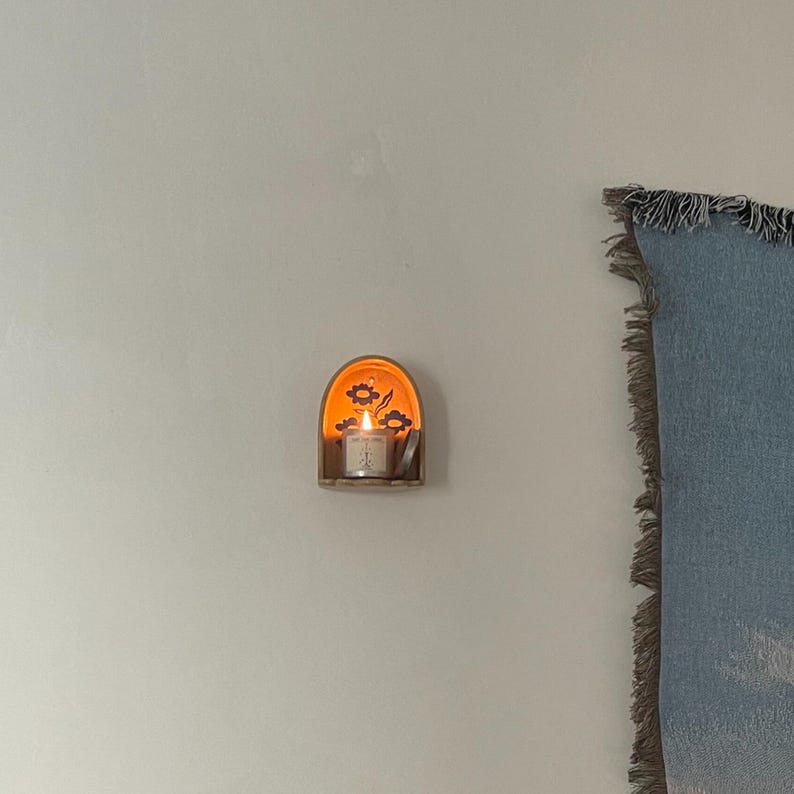 Pu&ograve; includere: Un piccolo portacandele da parete in legno con una candela accesa. Il portacandele presenta un design floreale e un bagliore caldo e invitante. La candela &egrave; in un piccolo supporto di vetro, creando un'atmosfera accogliente. Il portacandele &egrave; montato su una parete bianca.